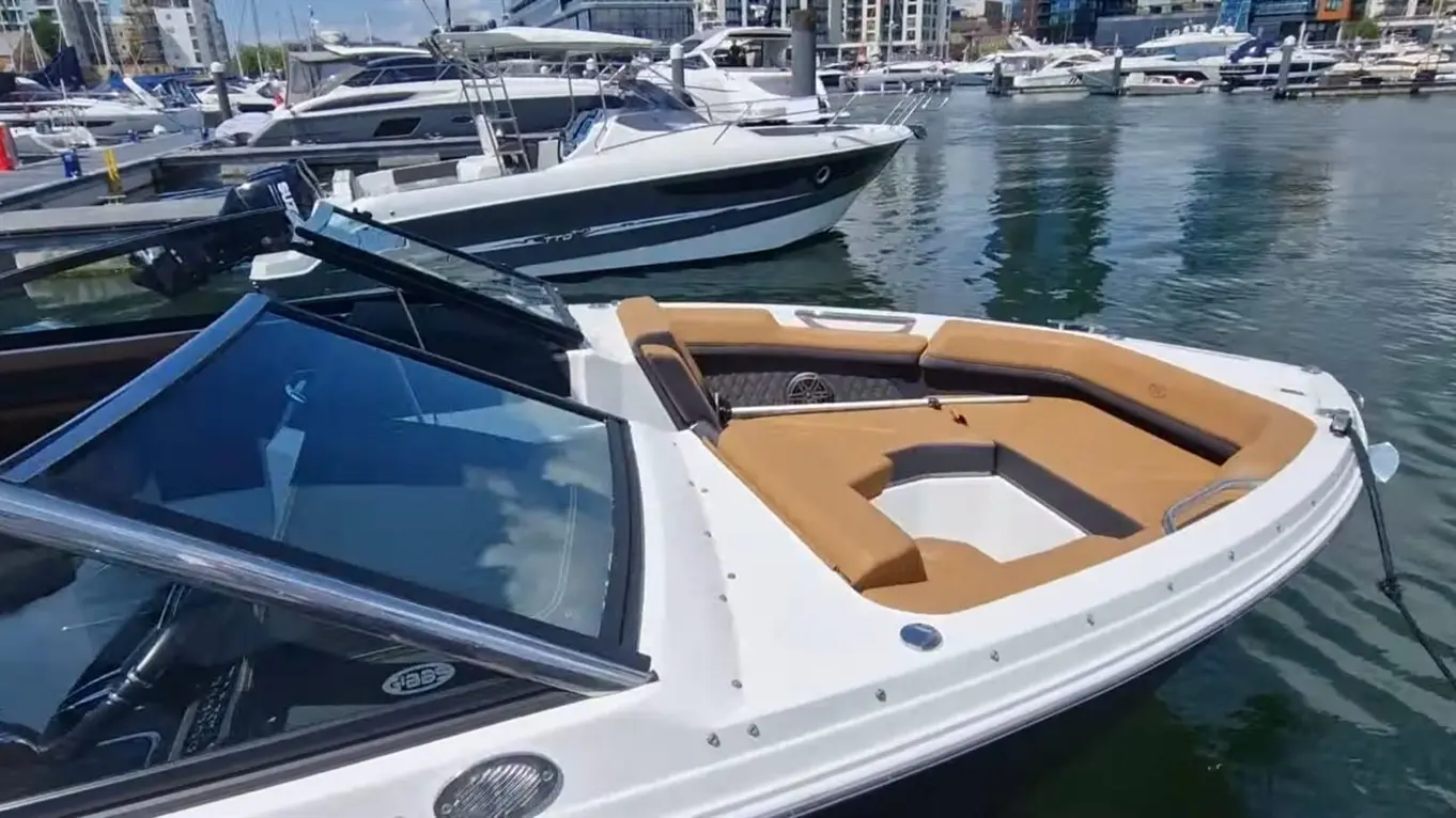 A sleek motorboat with a spacious tan seating area docked at a marina, surrounded by other boats on calm water.